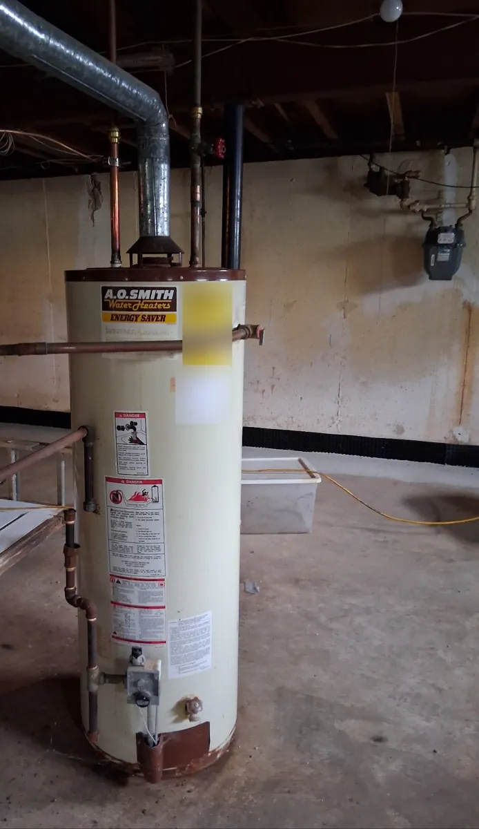 Old cream-colored gas tank water heater with a metal flue pipe rising to the ceiling in an unfinished basement
