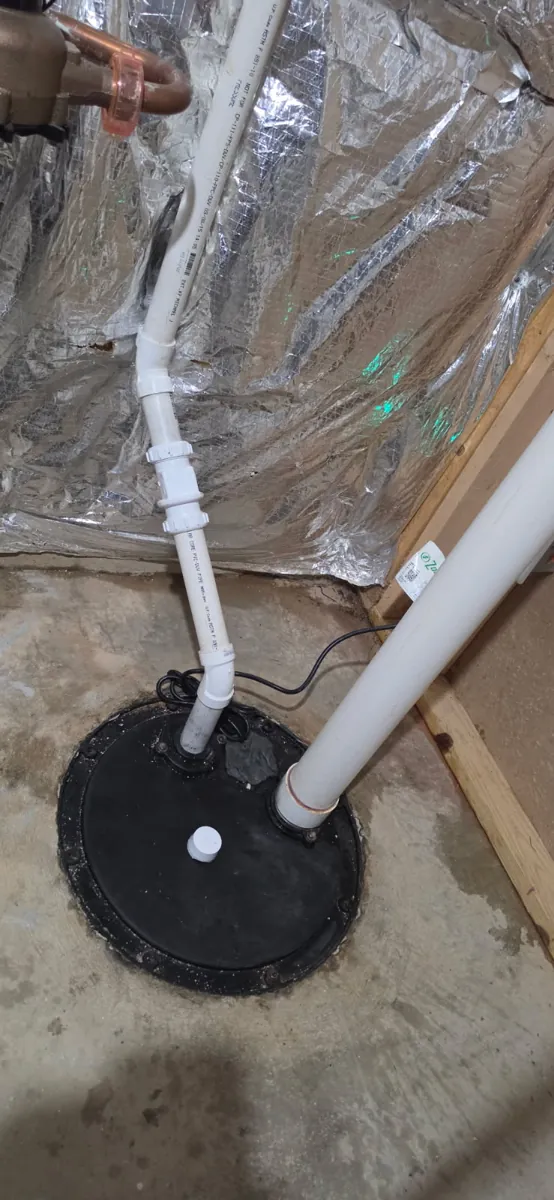 Finished sump pit with a sealed black lid, white PVC discharge pipe with unions rising from the pit, and a separate white PVC pipe
