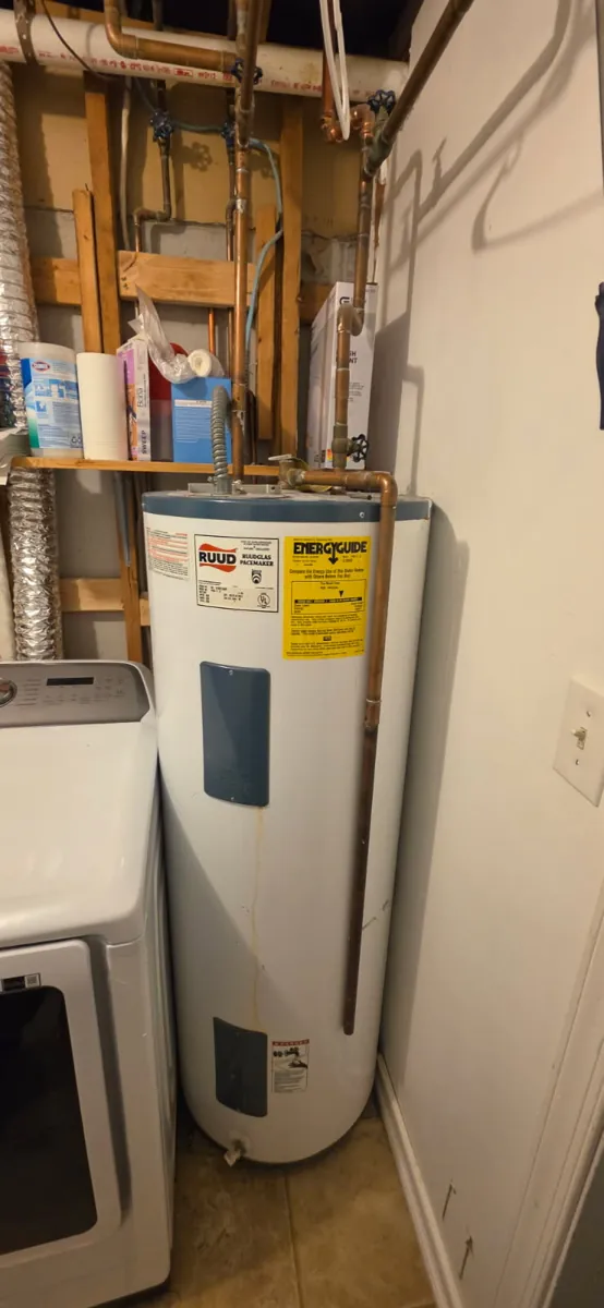 Old white Ruud electric water heater with a rust streak down the front, beside a dryer in a laundry room