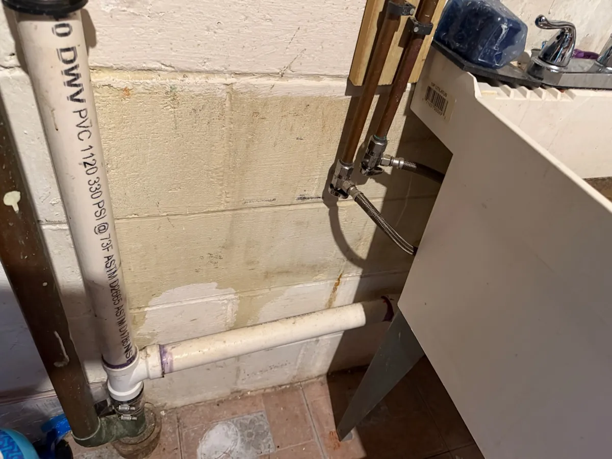 White PVC drain pipe from a laundry tub running into a vertical PVC drain stack against a block wall