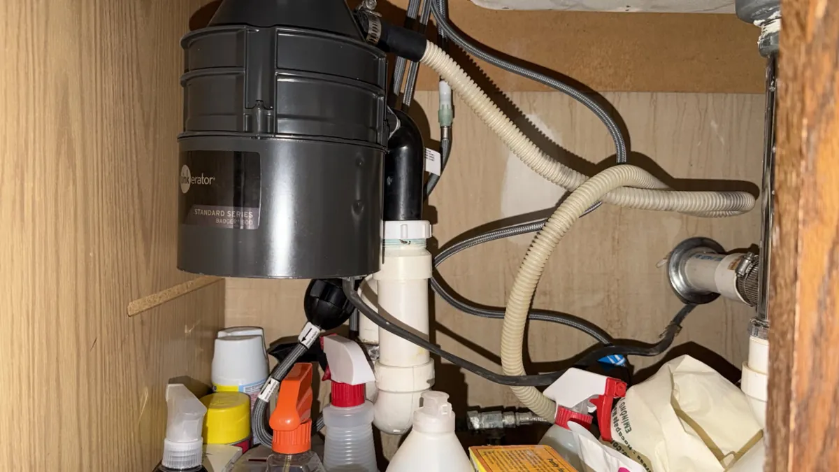 Under-sink cabinet with a black garbage disposal, white PVC drain pipes and a chrome tailpiece